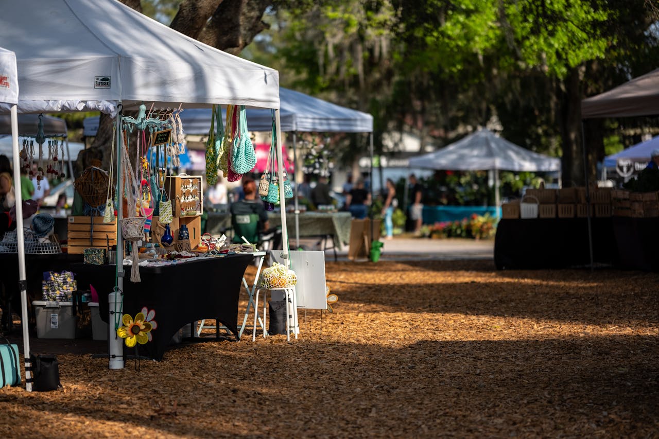 Vibrant outdoor market featuring craft stalls under tents with trees and people enjoying the atmosphere.