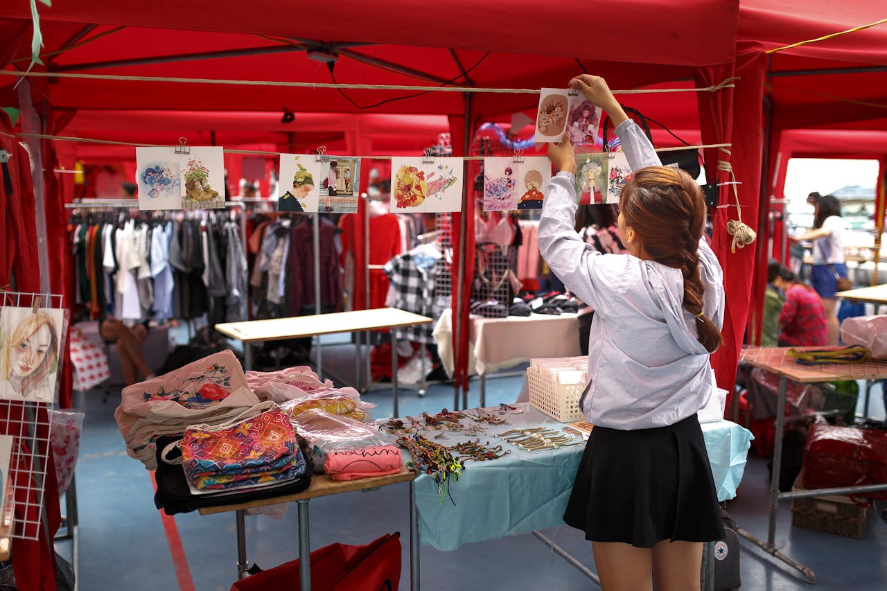 gallery-5 A woman organizes art prints in a vibrant outdoor market stall with colorful clothing and accessories.